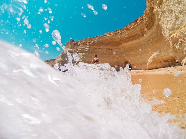 Portugal Urlaub. Welle bricht am Strand vor steilen Klippen in Portugal, mit einer Person am Fuß der Felsen unter klarem, blauem Himmel.