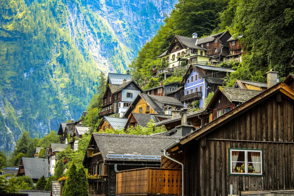 Urlaub Österreich. Traditionelles Bergdorf in den österreichischen Alpen mit Holzhäusern am Hang, umgeben von grünen Wäldern und steilen Felsformationen.