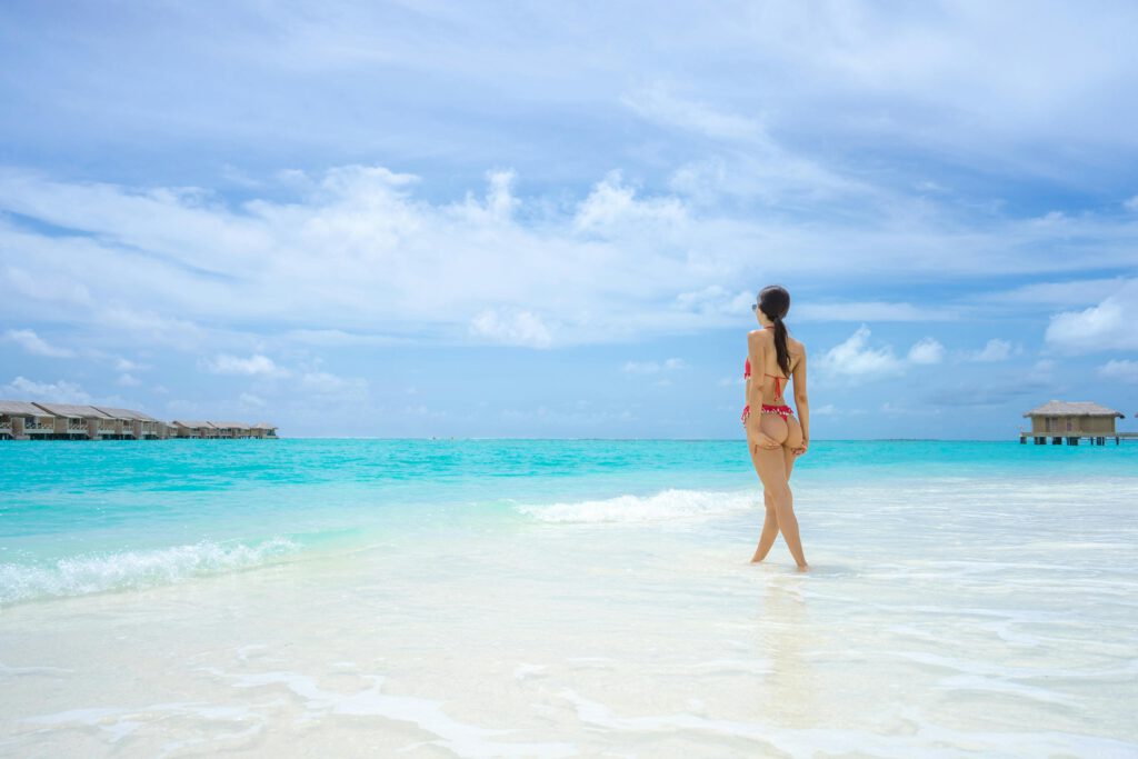 Frau läuft barfuß am weißen Sandstrand mit türkisfarbenem Wasser und Palmen im Hintergrund – Barfuß-Luxus auf den Malediven