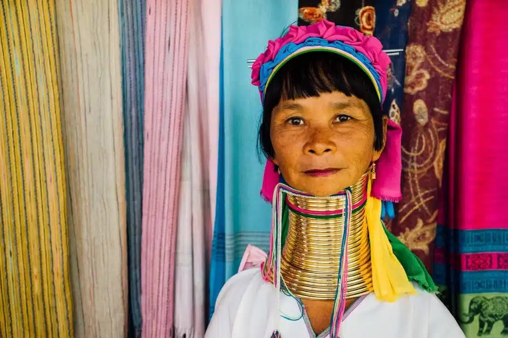 Nord Thailand Urlaub. Frau in traditioneller Kleidung mit buntem Kopfschmuck und Halsringen vor einem Marktstand mit farbenfrohen Stoffen – kulturelle Szene aus Nordthailand.