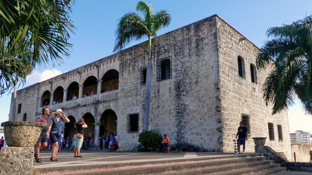 Urlaub Dominikanische Republik. Historisches Kolonialgebäude in Santo Domingo mit Touristen und Palmen bei strahlendem Himmel – Symbol für Kultur und Lebensfreude in der Dominikanischen Republik.
