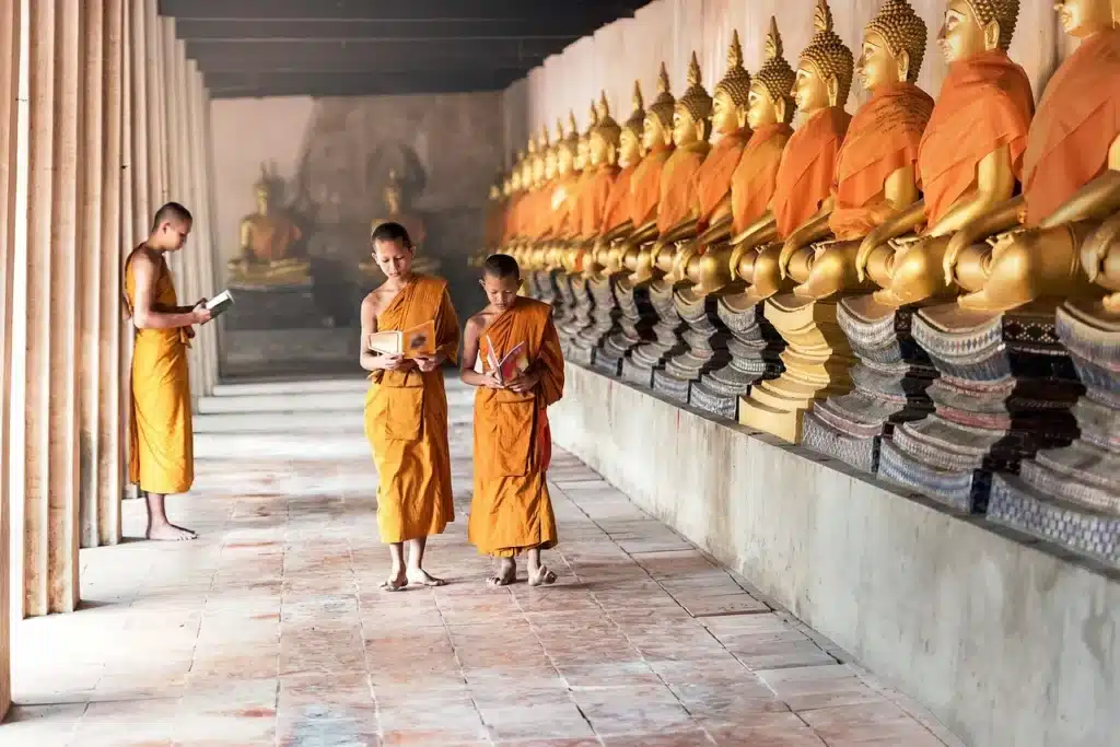 Drei Personen in orangefarbenen Gewändern gehen durch einen Tempelgang mit goldenen Buddha-Statuen – spirituelle Szene aus Thailand.