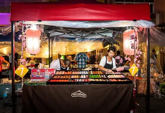 Phuket Urlaub. Streetfood-Stand mit rotem Dach und bunten Laternen in Phuket, Thailand – Koch bereitet lokale Spezialitäten in lebendiger Marktumgebung zu.
