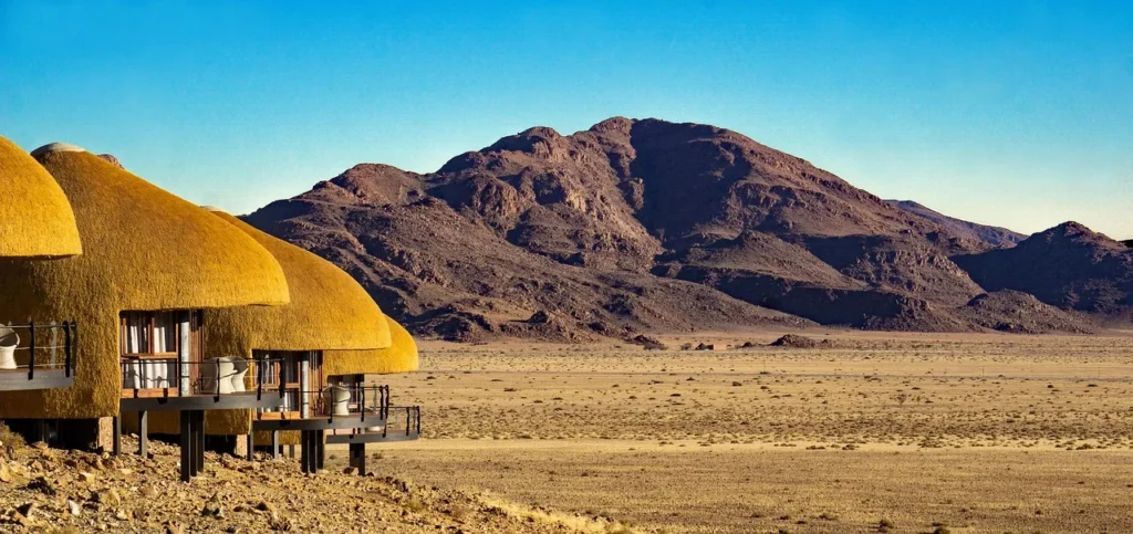 Rundhütten auf Stelzen in der Wüste vor Felskulisse – Desert Lodge in Namibia bei klarem Himmel