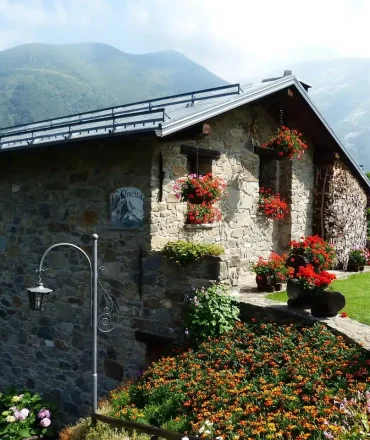 Steinhaus mit roten Blumenkästen, umgeben von blühendem Garten und grüner Landschaft, vor alpiner Bergkulisse.