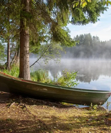 Natur Skandinavien. Holzboot am nebligen Seeufer, umgeben von Waldlandschaft in Skandinavien – ruhige Naturkulisse für nachhaltigen Urlaub.