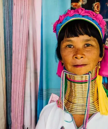 Nord Thailand Urlaub. Frau in traditioneller Kleidung mit buntem Kopfschmuck und Halsringen vor einem Marktstand mit farbenfrohen Stoffen – kulturelle Szene aus Nordthailand.