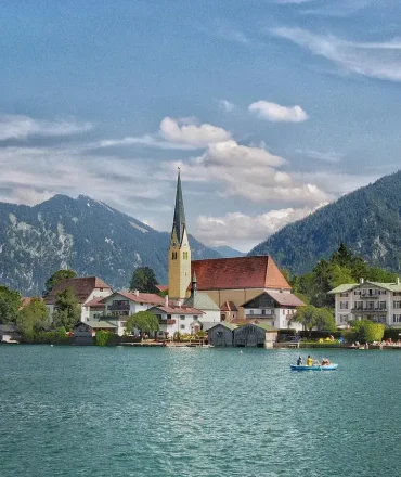 Urlaub Süddeutschland: Blick auf den Tegernsee mit Kirche, Alpenpanorama und Boot im Vordergrund