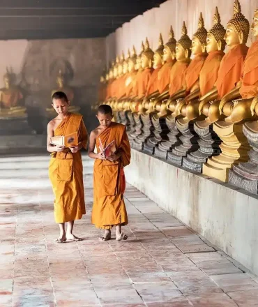Drei Personen in orangefarbenen Gewändern gehen durch einen Tempelgang mit goldenen Buddha-Statuen – spirituelle Szene aus Thailand.