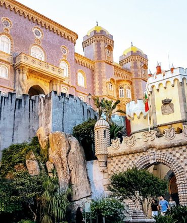 Last Minute Urlaub Portugal. Palácio da Pena in Portugal – farbenfrohe Fassade mit Türmen und Balkon über grüner Landschaft