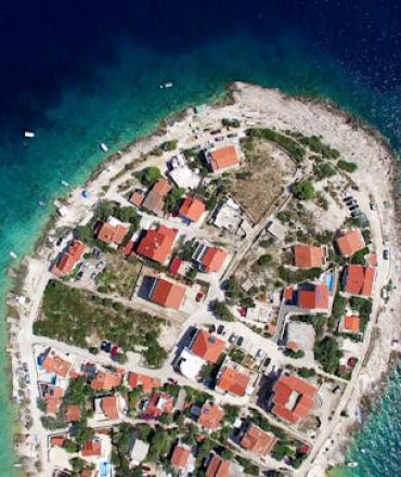 Küstenstadt mit roten Dächern auf schmaler Landzunge, umgeben von türkisblauem Wasser und grüner Landschaft