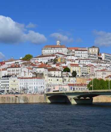 Portugal Urlaub. Panoramablick auf Coimbra mit der Brücke über den Mondego, historischen Häusern auf dem Hügel und teils bewölktem Himmel – Werbebild für Portugal All Inclusive Urlaub