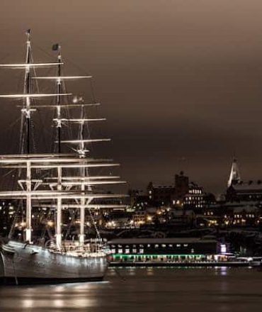 Schweden Urlaub. Beleuchtetes Segelschiff bei Nacht im Hafen von Stockholm, Schweden, mit Spiegelung im Wasser und Stadtpanorama im Hintergrund.