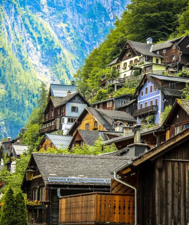 Urlaub Österreich. Traditionelles Bergdorf in den österreichischen Alpen mit Holzhäusern am Hang, umgeben von grünen Wäldern und steilen Felsformationen.