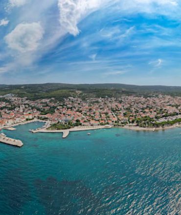 Last Minute Kroatien. Panorama einer kroatischen Küstenstadt mit roten Dächern, Hafen und türkisblauem Meer, umgeben von Natur