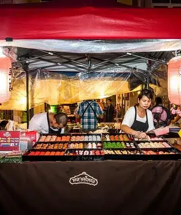Phuket Urlaub. Streetfood-Stand mit rotem Dach und bunten Laternen in Phuket, Thailand – Koch bereitet lokale Spezialitäten in lebendiger Marktumgebung zu.