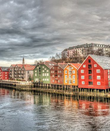 Bunte Häuser am Wasser in Trondheim, Norwegen, mit Spiegelung im Fjord und Brücke im Hintergrund – typisch für skandinavische Küstenstädte.
